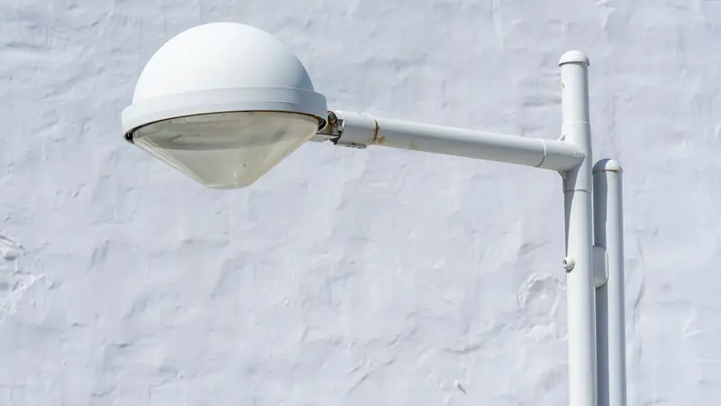 Luminaire extérieur blanc sur mât, fixé devant un mur blanc crépi, en plein jour