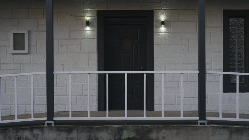 Porte d'entrée sombre encadrée de deux appliques murales allumées, sous un porche à garde-corps blanc