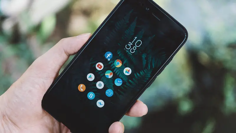 Main tenant un smartphone Android dont l'écran d'accueil affiche des icônes d'applications rondes sur un fond de fougères