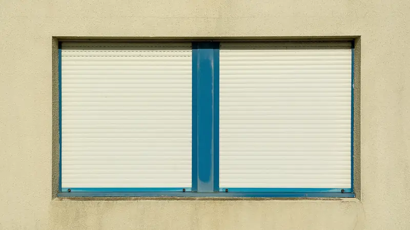 Fenêtre double encadrée de bleu sur un mur beige, ses deux volets roulants blancs entièrement fermés