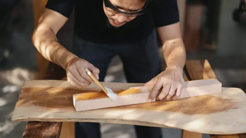 Artisan appliquant un vernis au pinceau sur une pièce de bois posée sur un tréteau