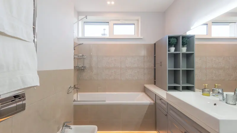 Salle de bain beige avec baignoire, pare-bain vitré, double vasque et sèche-serviettes chromé garni de serviettes blanches
