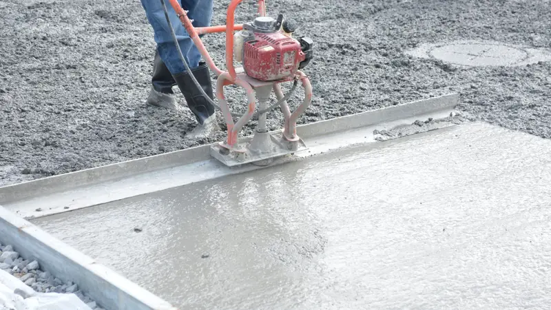 Ouvrier en bottes lissant une dalle de béton frais avec une règle vibrante thermique rouge posée sur des coffrages métalliques