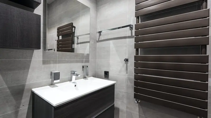 Radiateur sèche-serviettes à lames brunes fixé au mur d'une salle de bains carrelée de gris, à côté d'un meuble vasque